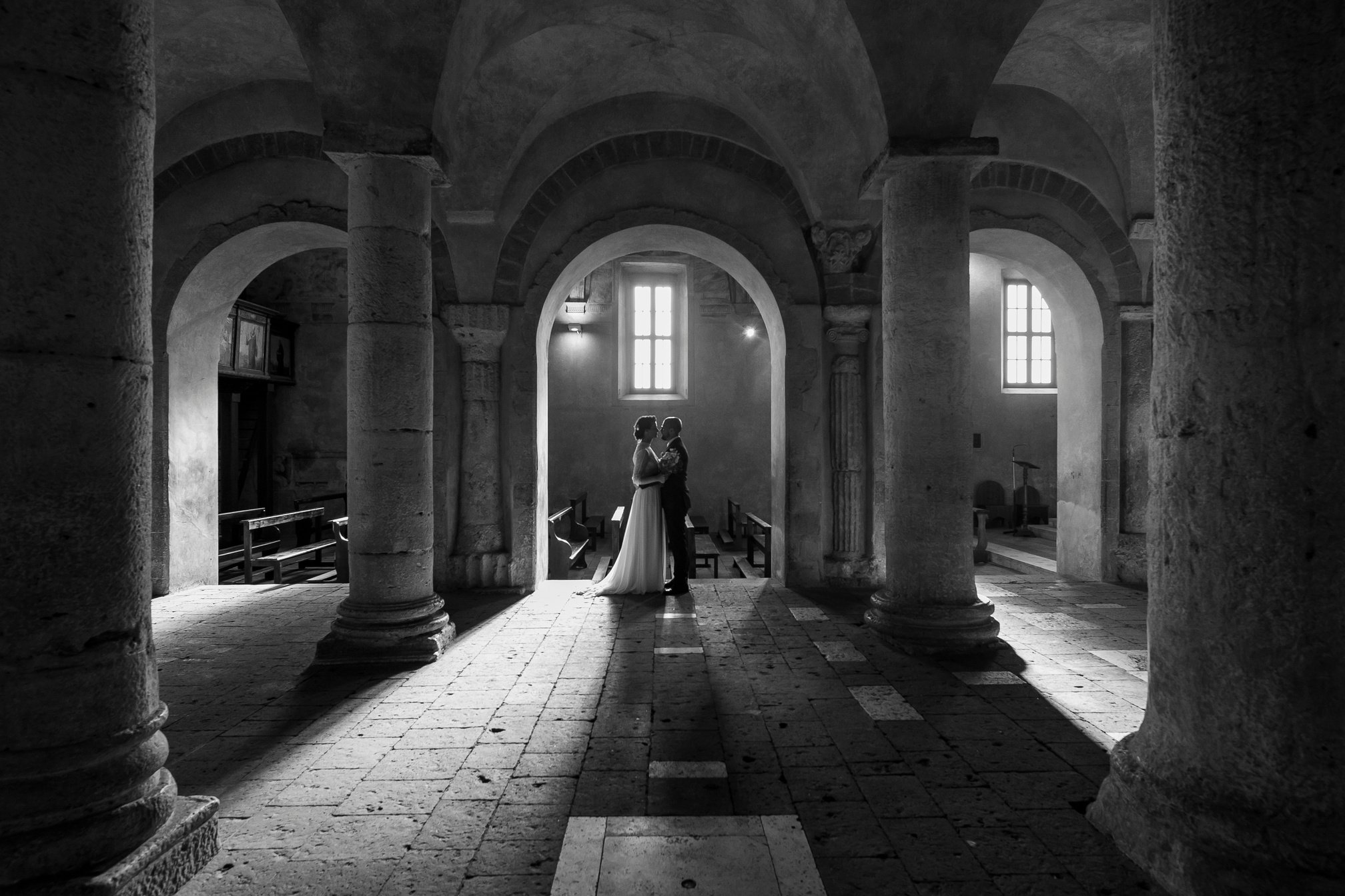 Sotto gli archi di una chiesa a Cori, la foto ritrae la coppia di sposi. Il fotografo matrimonio cattura la solennità della cerimonia, creando un'immagine emozionale. Servizio fotografico matrimonio Cori